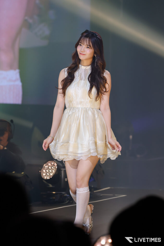 Female performer in a cream lace dress with long wavy hair on stage, smiling at the audience. Full-body shot with stage lights in the background.