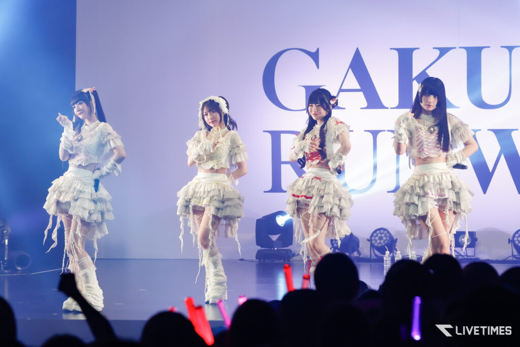Four female performers in white, frilly costumes dance on a lit stage with a large backdrop of letters behind them, blue lighting.