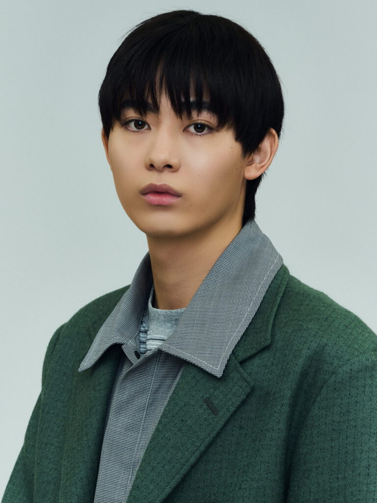 Close-up portrait of a young person with short black hair, wearing a green coat over a gray checkered shirt, facing the camera.