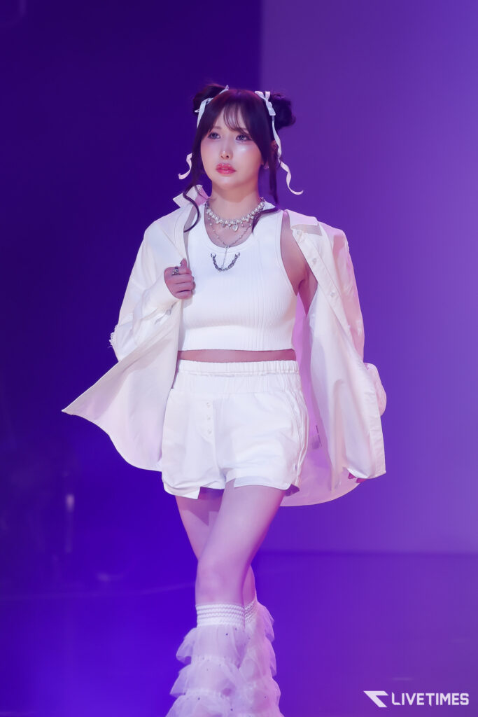 Young woman performing on stage in a white crop top and jacket, purple lighting in the background, wearing layered necklaces.