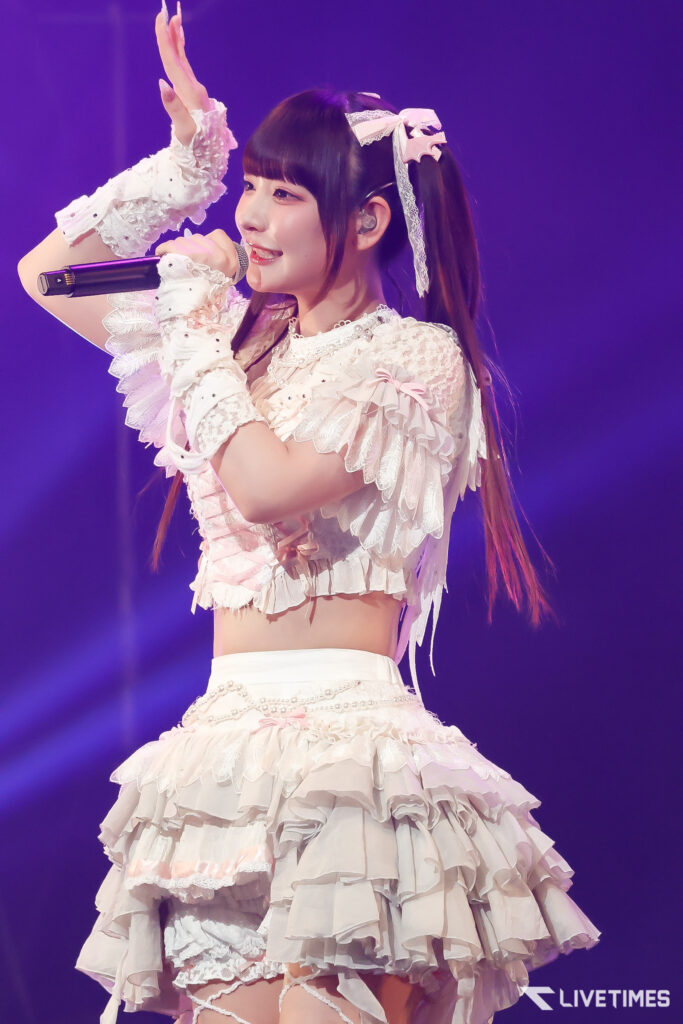 Female singer on stage wearing a white frilly dress, holding a microphone and raising her hand in a performance pose.