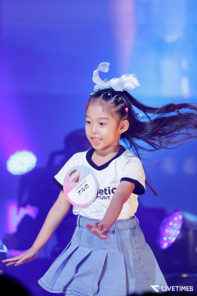 Young girl dancing on stage with a white bow in her hair and a circular badge on her shirt, blue lighting in the background