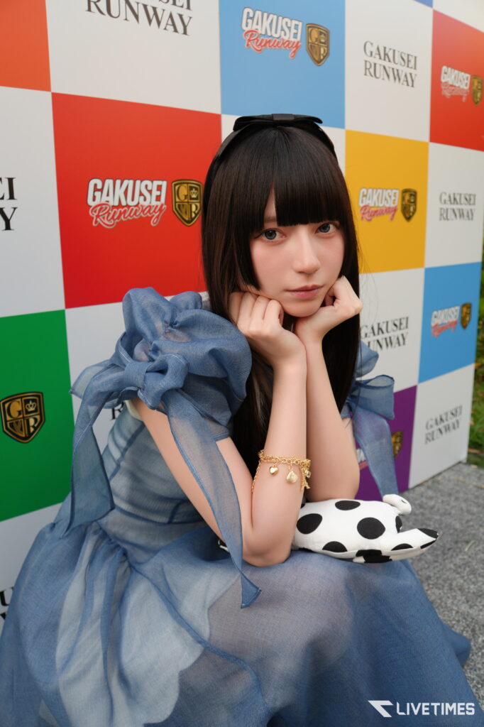 Young woman with long black hair in a blue dress posing with hands under her chin in front of a colorful checkered backdrop with logos, including Gakusei Runway