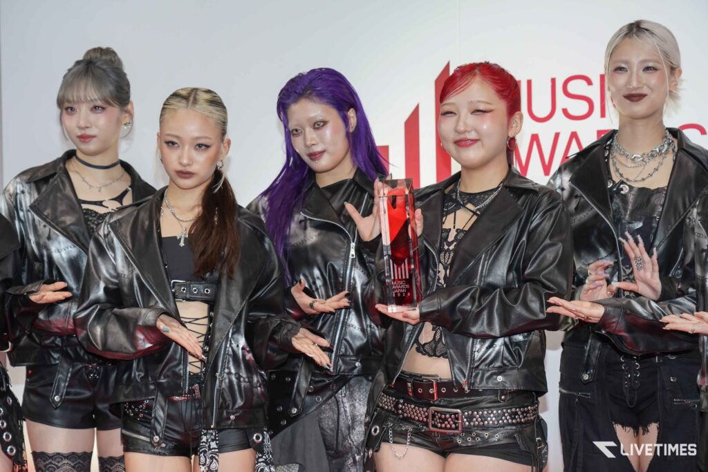 Group of five women in black leather outfits posing with a tall red trophy at a music awards event (visible 'MUSIC AWARDS JAPAN 2026').
