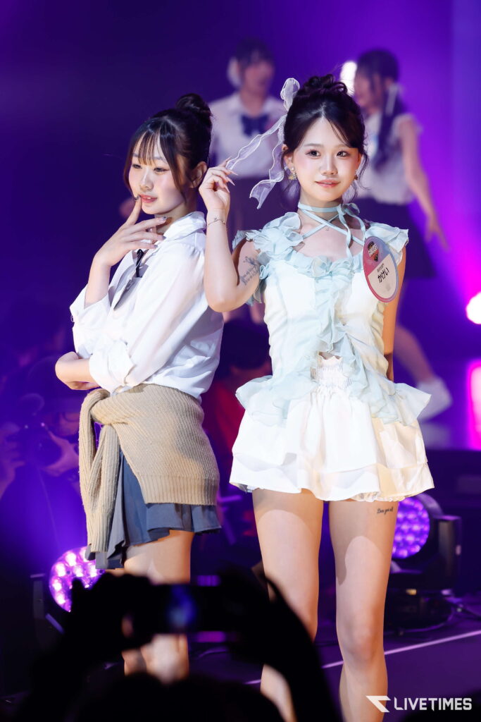 Two young performers on stage under purple lights, posing back-to-back as fans film them in the foreground. Two female performers strike poses during a concert, vibrant stage lighting behind them.