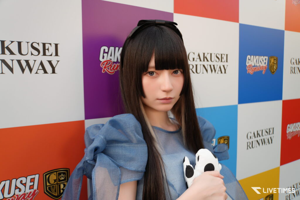 Young woman with long dark hair and bangs posing in front of a colorful GAKUSEI RUNWAY backdrop, holding a panda plush."