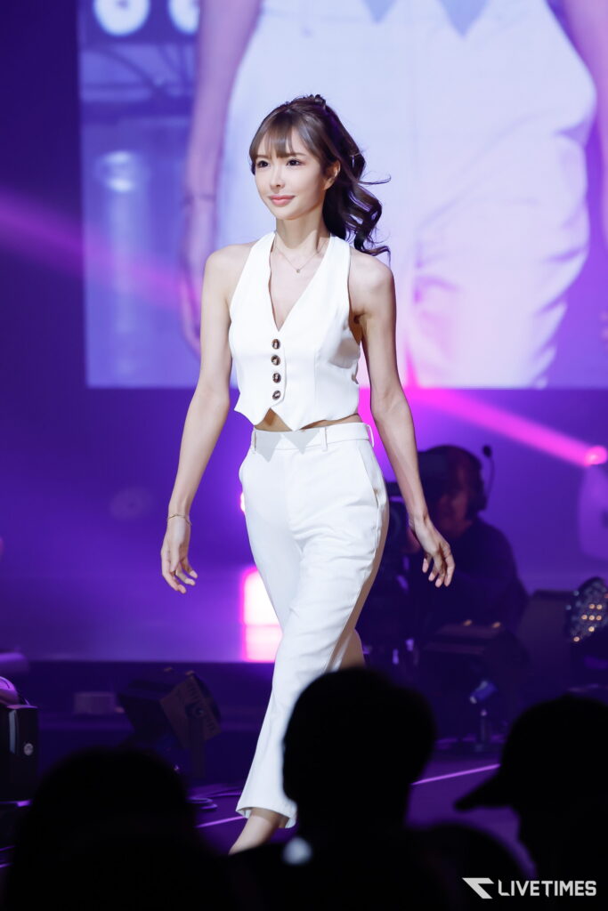 Female performer in a white sleeveless outfit walking on a purple-lit stage with audience silhouettes in the foreground.