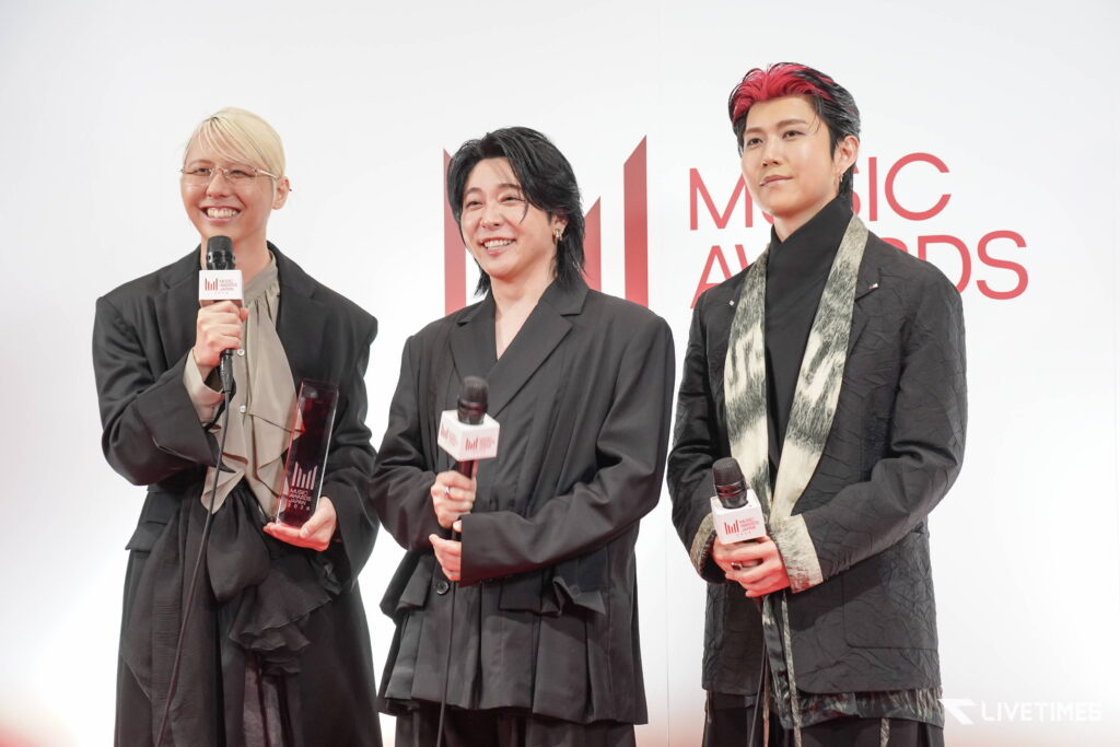 Three award winners stand on a white stage, each holding a microphone; they wear black outfits and smile for the camera.