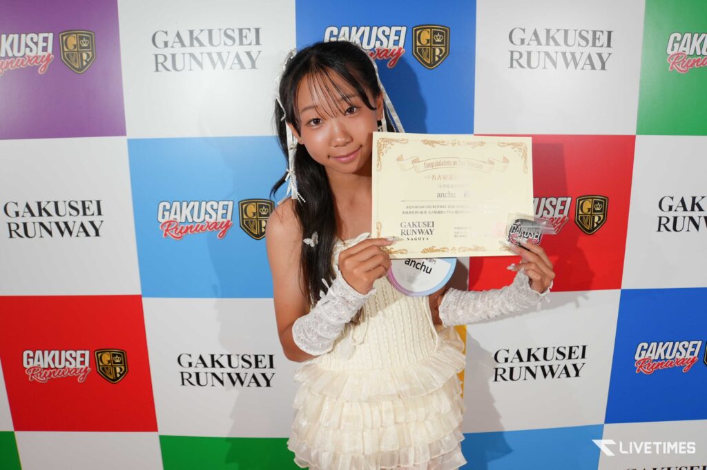 Smiling young woman in a white, frilly dress holds a certificate and medal at a colorful backdrop that says Gakusei Runway and Nagoya