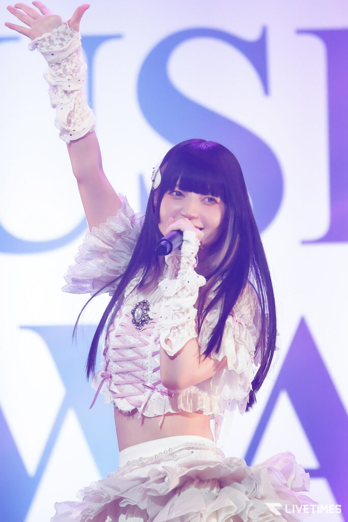 Young female singer on stage raises her right arm in a frilly white costume while singing into a microphone.