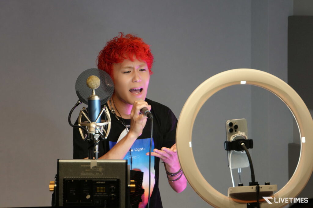 Singer with bright red hair performing into a microphone in a recording studio, with a pop filter and ring light nearby.