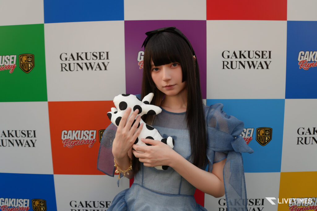 Portrait of a young woman with long black hair, holding a black‑polka‑dot plush toy, standing in front of a colorful GAKUSEI RUNWAY backdrop.