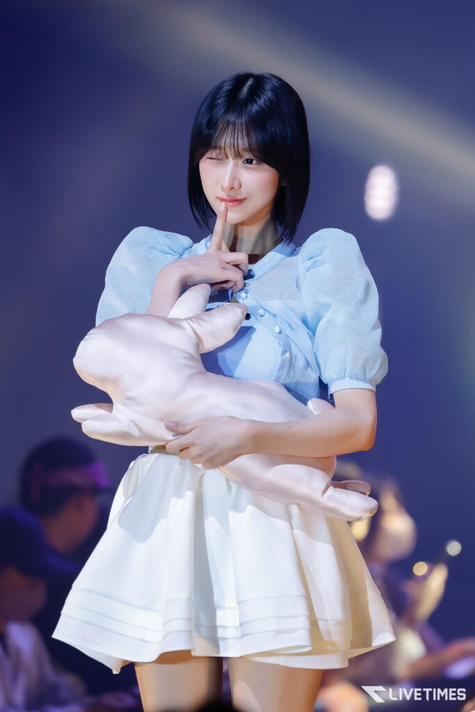 Woman on stage winking and touching her lips with her finger, wearing a light blue puff-sleeve top and white skirt.
