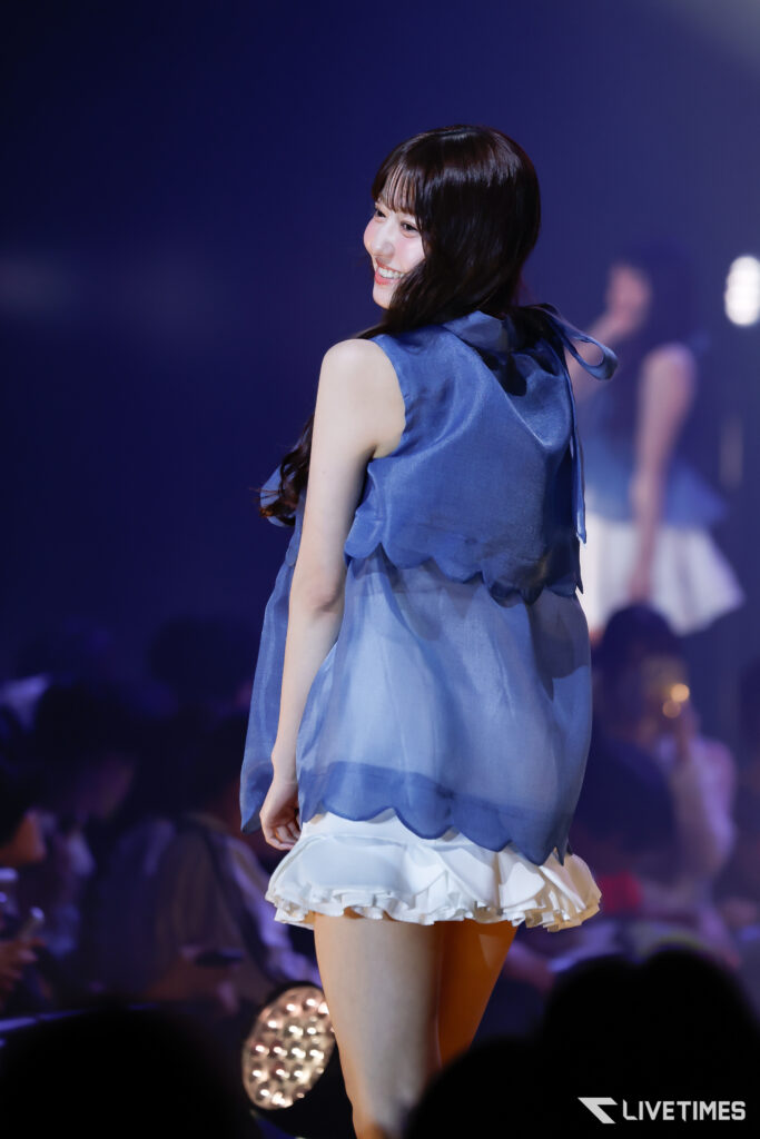 Woman on stage in a blue sleeveless dress, smiling as she turns to the side; soft stage lighting and blurred audience in the background.