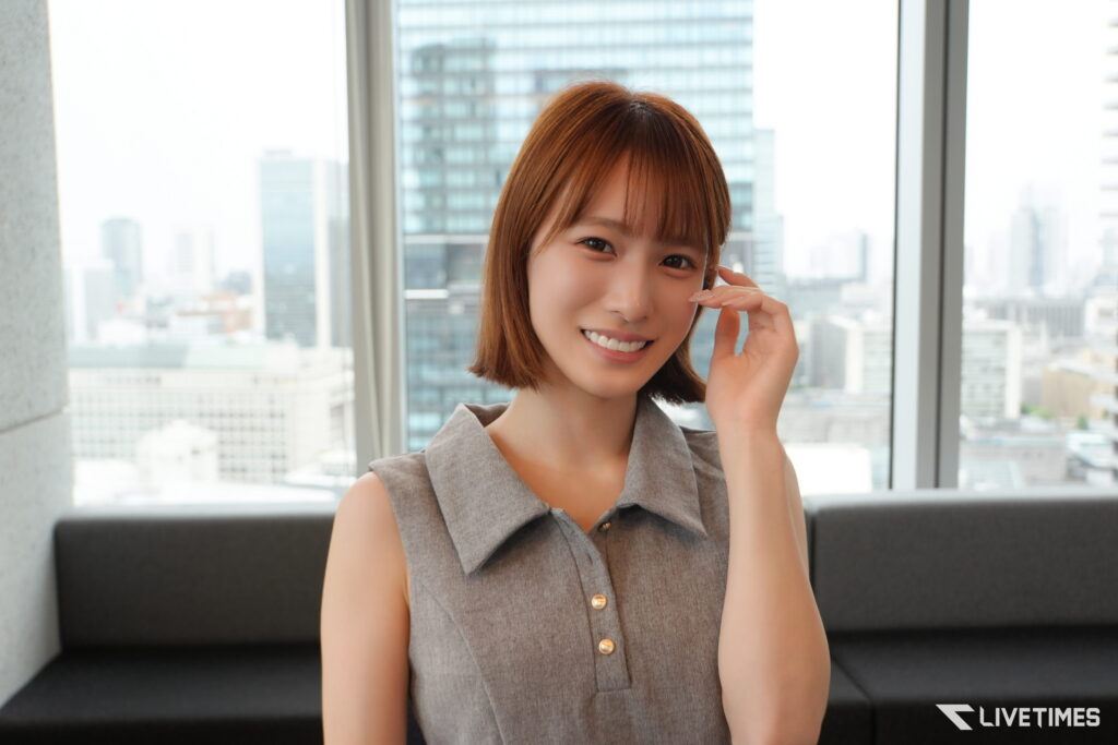 Smiling woman with short hair in a sleeveless gray dress sits indoors with a city skyline outside the window behind her.