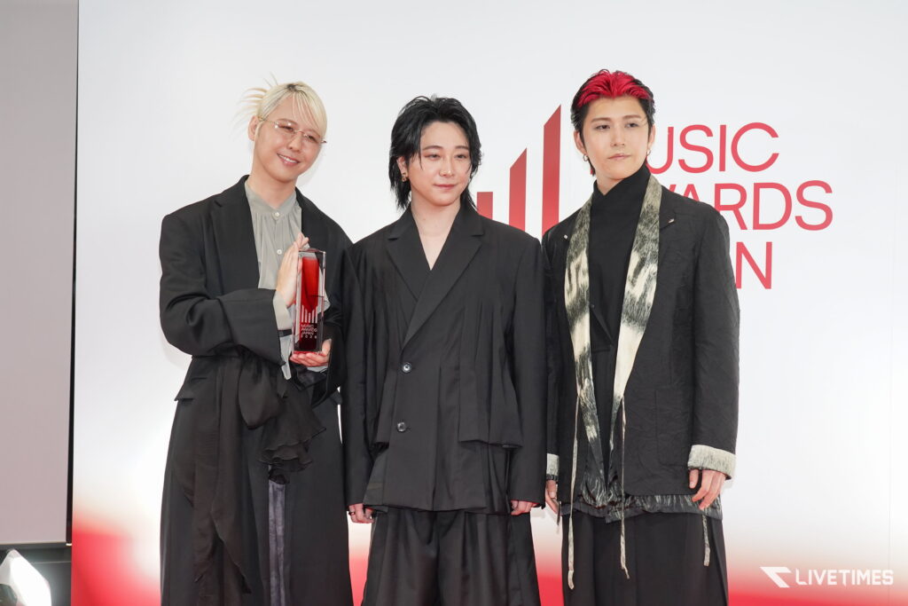 Three people in dark outfits pose together on a red-carpet backdrop at a music awards event; the person on the left holds a red trophy and smiles softly while wearing light-colored glasses.