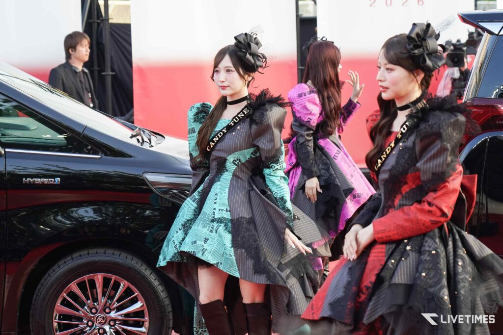 Three models in colorful, patterned outfits walk beside a black car at a red-carpet event, one wearing a black fascinator with netting and a sash reading CANDY TUNE.