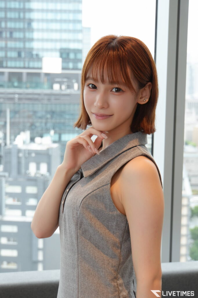Portrait of a woman with short brown hair in a gray sleeveless dress, posing by a window in a modern office building and smiling at the camera.