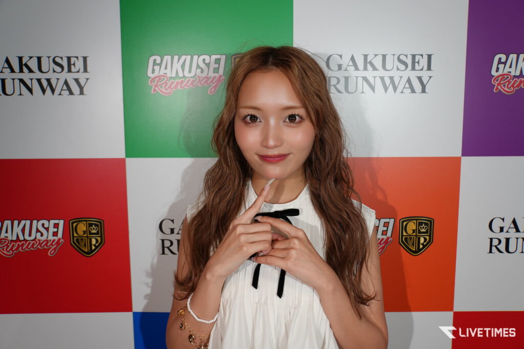 Young woman with wavy hair in a white blouse poses with her finger on her chin in front of a colorful backdrop with logos.