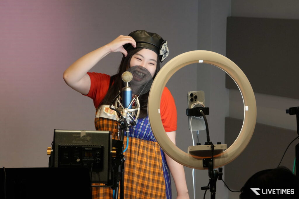 Person recording in a studio with a microphone, pop filter, and ring light; wearing a black cap and colorful outfit.