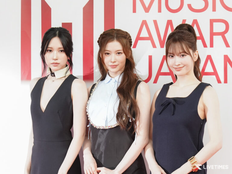 Three women in formal dresses pose for photos in front of a red-and-white backdrop at a music awards event.