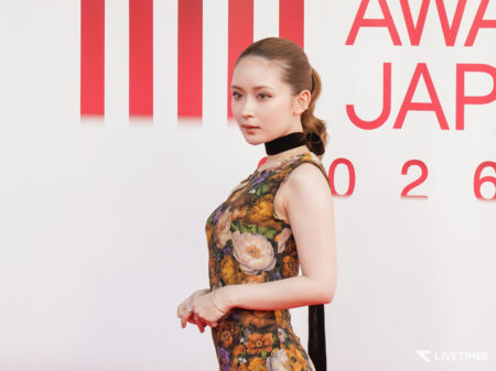 Side profile of a woman in a floral dress with a black choker, standing against a white wall with red vertical stripes and partial red text in the background.