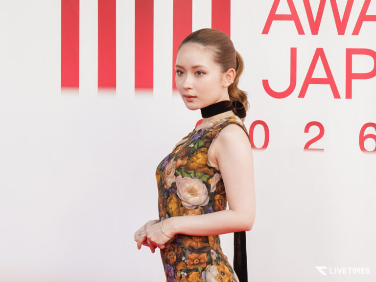 Side profile of a woman in a floral dress with a black choker, standing against a white wall with red vertical stripes and partial red text in the background.
