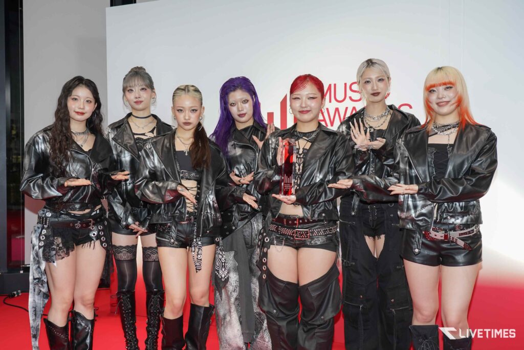 Group of seven women in black leather outfits posing on a red carpet at a music awards event, one holding a clear trophy with red accents.