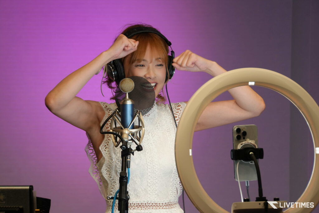 Female singer in headphones adjusts to recording mic in a studio with a purple wall behind her and a pop filter nearby and white lace top outfit.