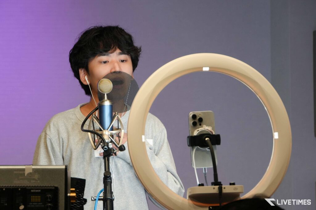 Person recording into a microphone with a pop filter in a studio, ring light visible nearby.