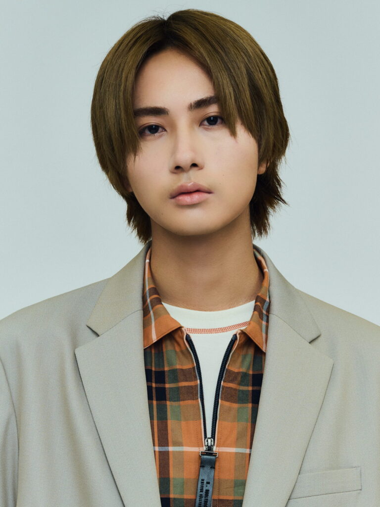 Portrait of a person with short brown hair wearing a beige blazer over an orange plaid shirt, facing forward against a pale background.
