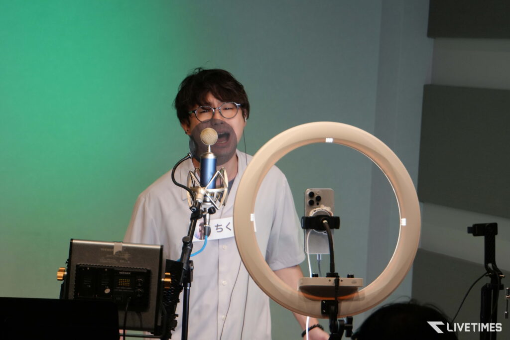 Person in a white lab coat and glasses speaks into a microphone with a pop filter in a recording studio, a circular ring light nearby and audio gear visible in the foreground.
