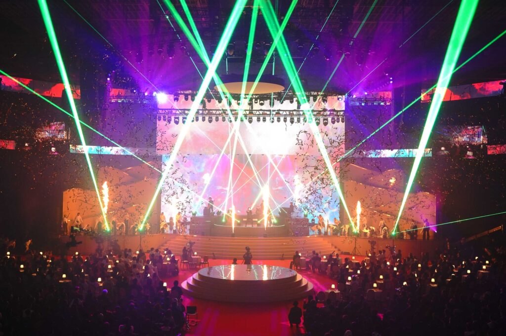 Concert stage lit by vivid green laser beams crossing over a multi-level set with a bright LED backdrop; audience seated at tables in a dark venue.