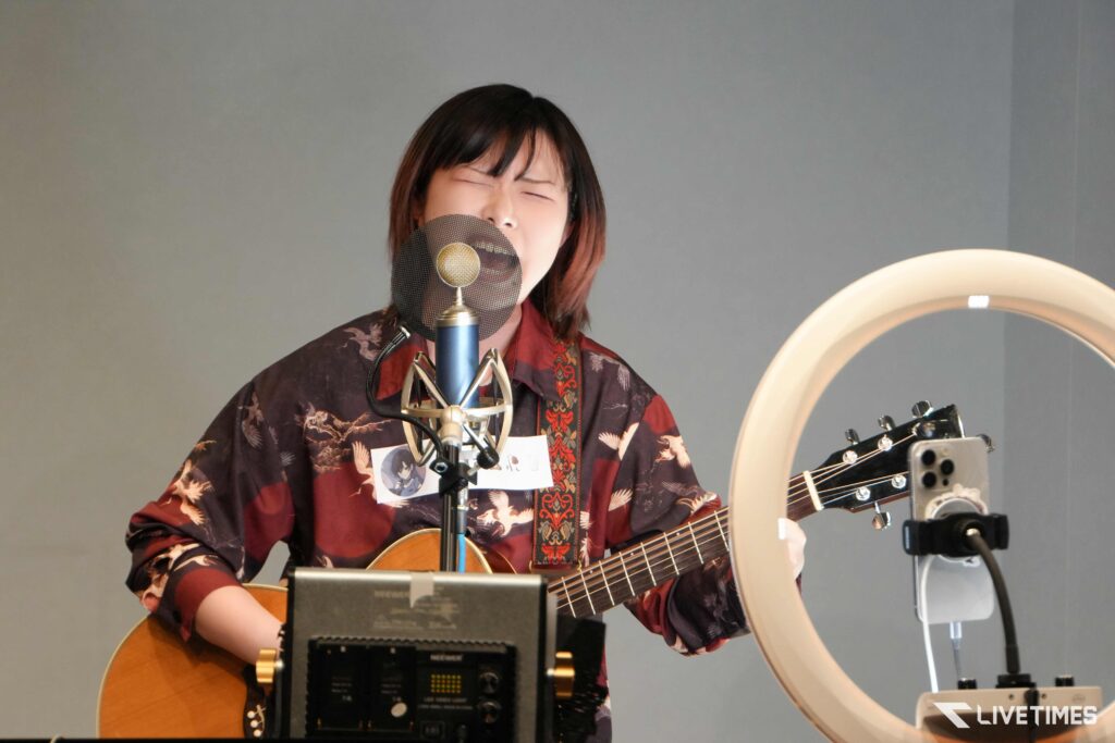 Female musician singing into a microphone with a pop filter while playing an acoustic guitar in a recording setup with a ring light nearby.