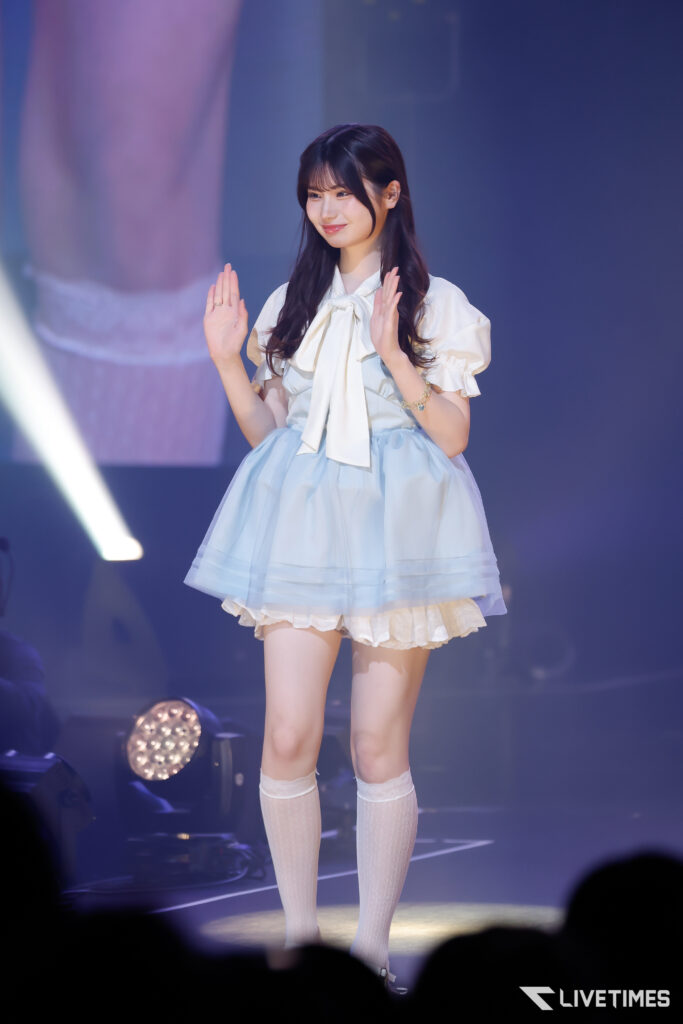 Young woman on stage in a pastel blue and white dress with a bow, waving to the audience under stage lights.