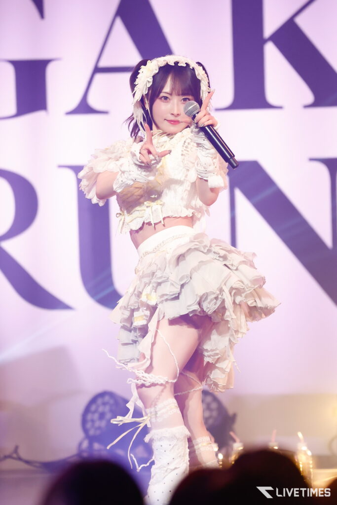 Young female singer in a frilly white stage costume with flower headband, holding a microphone and gesturing toward the camera on stage.