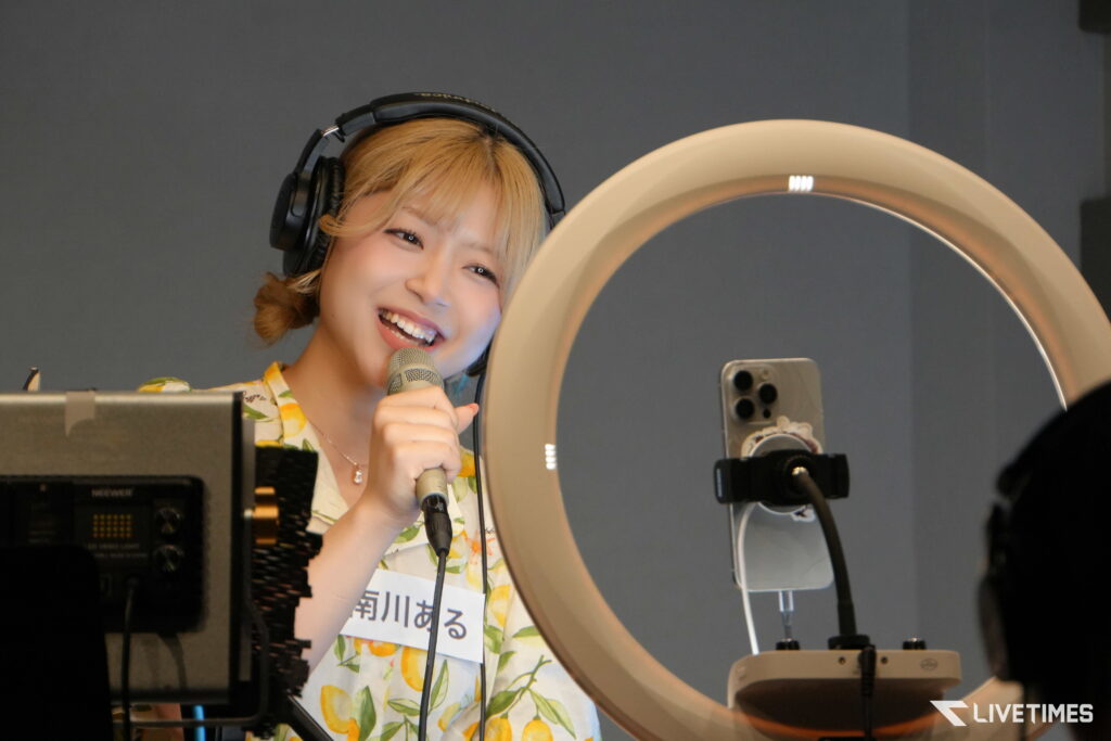 Woman wearing large over-ear headphones sings into a handheld microphone in a recording studio, smiling warmly at the mic beside a circular ring light.