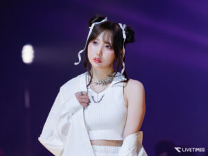 Close-up of a female performer on stage wearing a white outfit and pearl necklace, with purple stage lighting in the background.