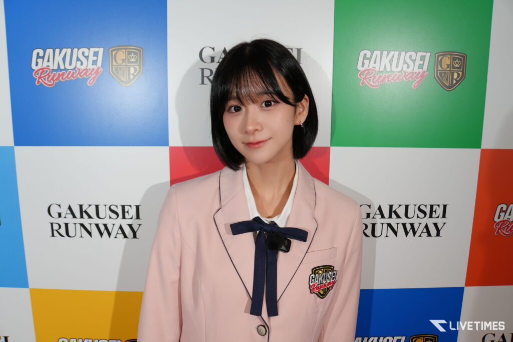 Portrait of a young woman in a pale pink blazer with a navy bow, standing before a colorful "GAKUSEI RUNWAY" backdrop.