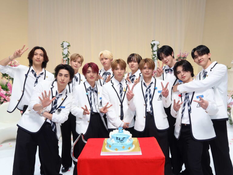 A group of smiling young men in matching white blazers and plaid ties pose around a birthday cake on a red table, making peace signs.