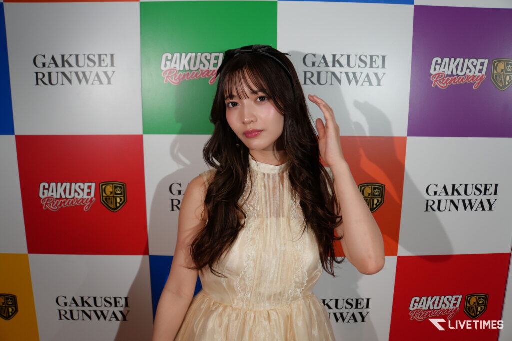 Young woman posing in a cream lace dress at a GAKUSEI RUNWAY backdrop with color blocks of red, green, blue, and white.