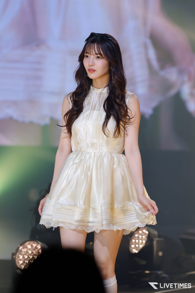 Young woman in a cream lace dress on stage, performing with soft lighting in the background.