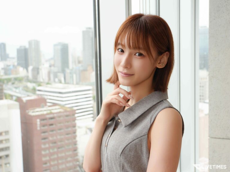 Woman with short brown hair in a gray sleeveless top stands by a window, city skyline visible outside and soft natural light.