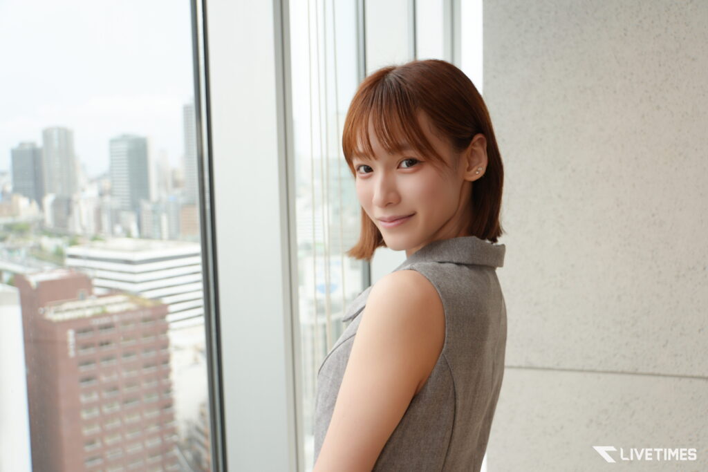 Woman with short brown hair in a gray sleeveless top standing by a large window, cityscape visible outside the glass panes.