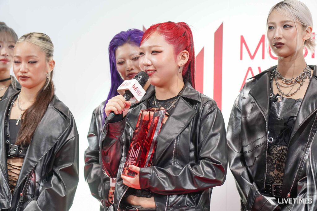 Group of young women in black leather outfits at an awards event; one woman with red hair speaks into a microphone and holds a glass trophy.
