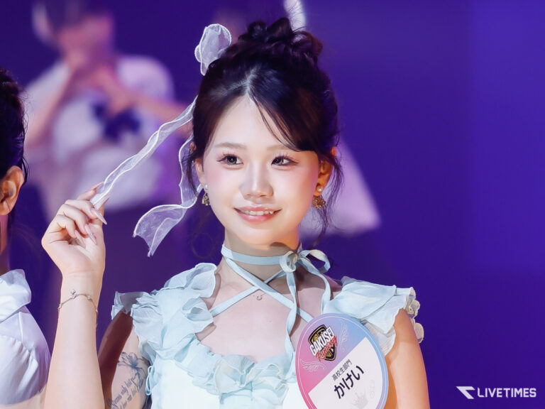 Young woman in a pale blue ruffled dress on stage, holding a sheer veil and smiling softly.