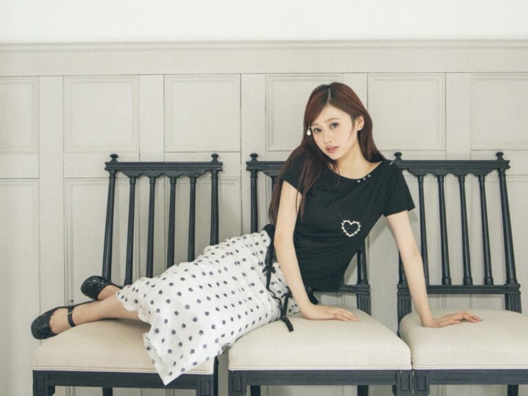 Young woman seated on a beige couch against a paneled wall, wearing a black top and white patterned skirt, looking at the camera.
