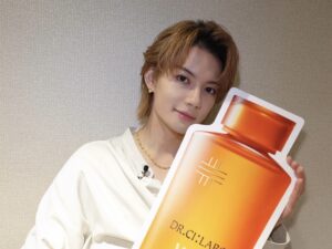 Person with light brown hair in a white shirt holds a large orange gradient brochure toward the camera, standing against a textured wall.