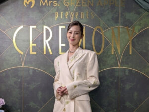 Woman in a cream suit with floral embroidery posing in front of a gold-letter backdrop that reads 'CEREMONY' at an event