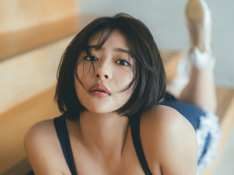 Close-up of a young woman with short black hair lying on wooden steps, looking at the camera, wearing a navy tank top and denim shorts visible in the background.